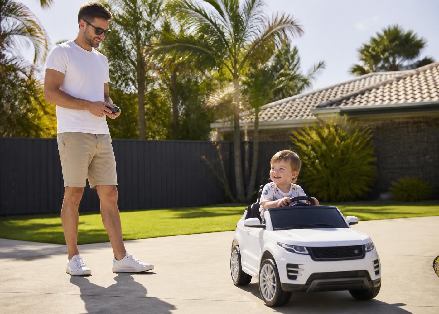 Parent using a parental remote control while a toddler drives a white kids ride on car